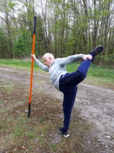 Mann hebt das hintere Bein im Stand und hält es mit der Hand, abgestützt mit Stab